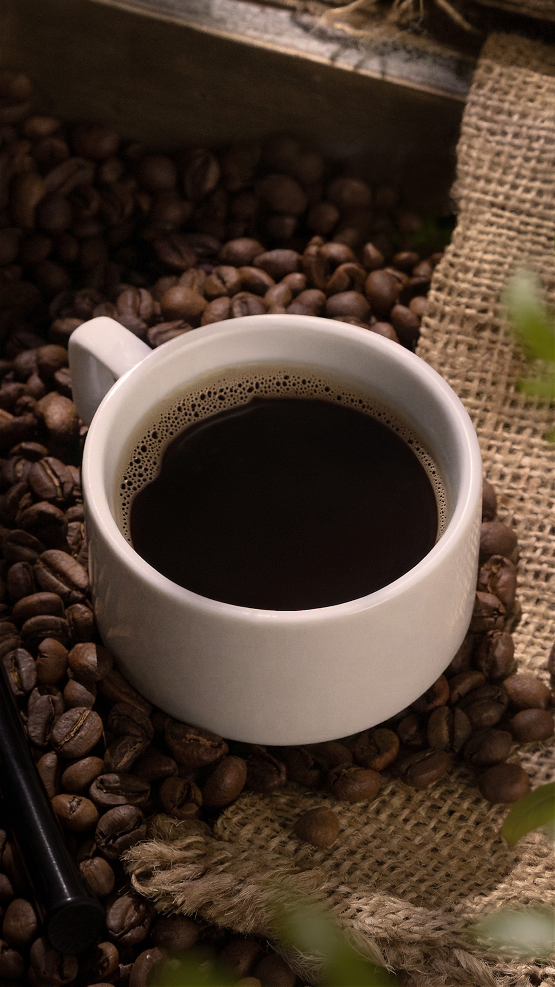 A white cup of black coffee on a bed of roasted coffee beans and burlap beside a pipe stem